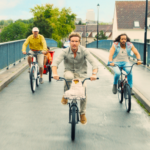 Trois hommes roulent rapidement à vélo sur un pont urbain sous un ciel nuageux