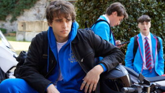 James assis sur une moto devant une école internationale à Milan dans Love Me Love Me sur Prime Video