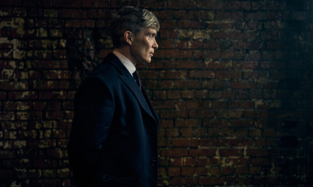 Tommy Shelby en costume sombre devant un mur de briques dans Peaky Blinders