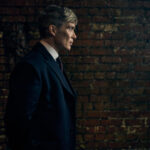 Tommy Shelby en costume sombre devant un mur de briques dans Peaky Blinders
