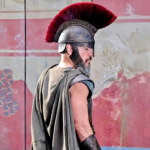 Soldat grec en armure avec casque à crête rouge pendant la guerre de Troie dans L’Odyssée de Christopher Nolan