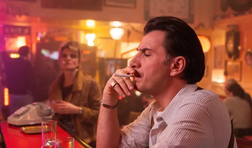Michaël Youn fumant une cigarette dans un bar à l’ambiance rétro des années 80, assis au comptoir avec un verre, tandis qu’une femme floue se tient en arrière-plan dans la série Flashback diffusée sur TF1
